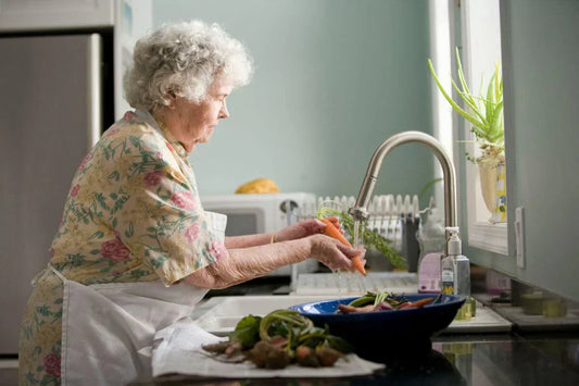 Elderly woman washing carrots at kitchen sink for an antioxidant-rich diet and cellular health.